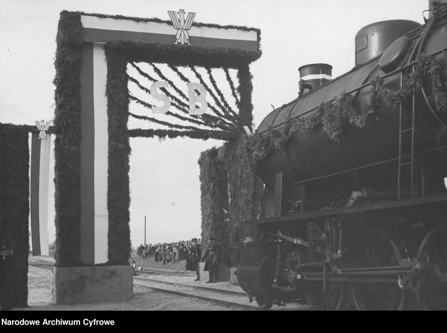Uroczysty wjazd pierwszego pociągu przez bramę triumfalną. Widoczny udekorowany parowóz Ty 23. Ze zbiorów Narodowego Archiwum Cyfrowego